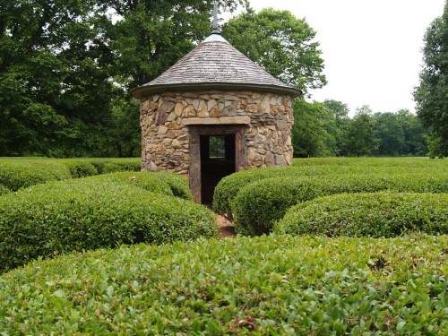 A stone gazebo surrounded by neatly trimmed green hedges in a lush garden setting.