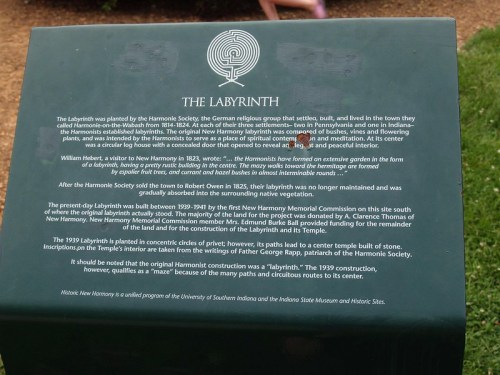 A sign about the Labyrinth, detailing its history and significance in New Harmony, Indiana, with a garden backdrop.