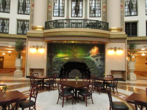 Elegant lobby with a mural, tables, and chairs, featuring decorative columns and lush greenery.