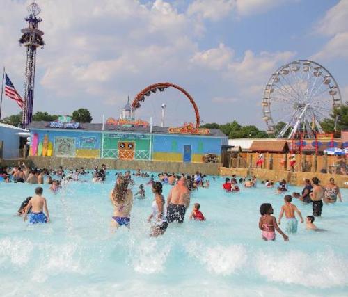 A crowded water park with people enjoying the wave pool, rides, and a ferris wheel in the background under a blue sky.