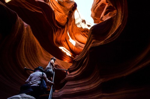 Visitors ascend a staircase through the stunning, wavy rock formations of Antelope Canyon, illuminated by sunlight.