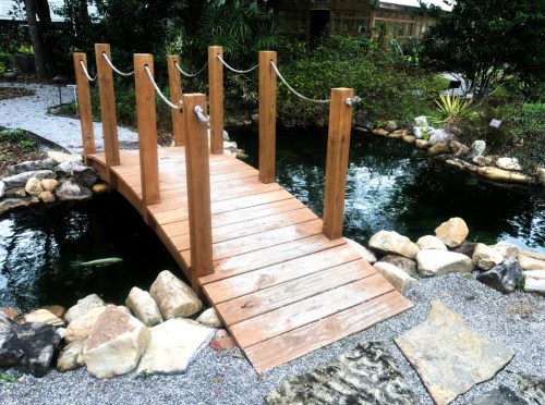 A wooden bridge with rope railings spans a small pond surrounded by rocks and greenery.