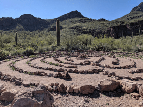 A stone labyrinth set in a desert landscape, surrounded by cacti and mountains under a clear blue sky.