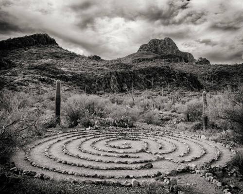 A black and white image of a stone labyrinth in a desert landscape, surrounded by mountains and cacti under a cloudy sky.