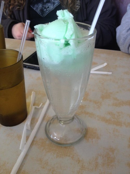 A tall glass filled with clear soda and topped with a scoop of green mint ice cream, with straws beside it.