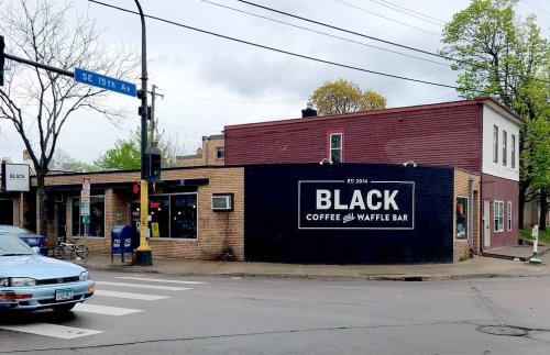 A corner building featuring a sign that reads "BLACK COFFEE & WAFFLE BAR," with trees and a street sign nearby.