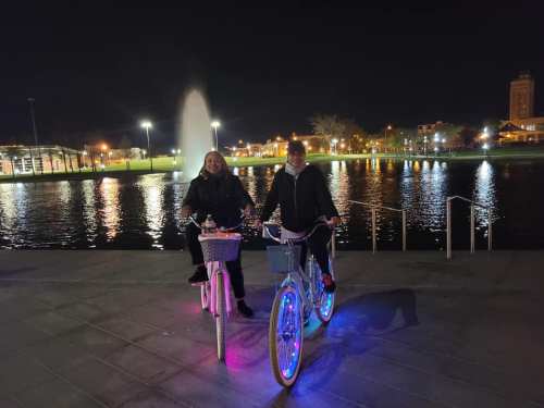 Two people on illuminated bicycles by a fountain at night, with city lights reflecting on the water.
