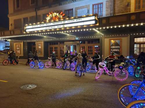 A group of people with brightly lit bicycles gathered outside a theater at night, under a marquee sign.
