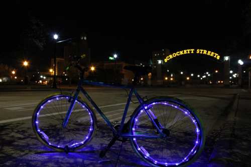 A blue bicycle with glowing wheels parked at night under a lit archway that reads "Crockett Street."