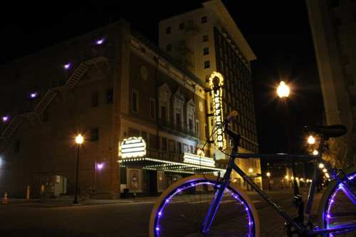 A blue bicycle with glowing wheels parked near a historic theater at night, illuminated by streetlights.
