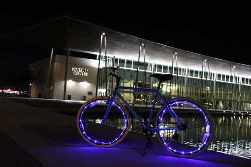 A brightly lit bicycle with glowing wheels parked near a modern building at night, reflecting in the water.
