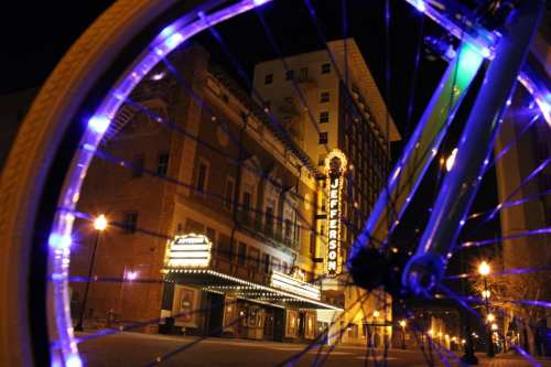A brightly lit bicycle wheel in the foreground with the Jefferson Theater illuminated at night in the background.