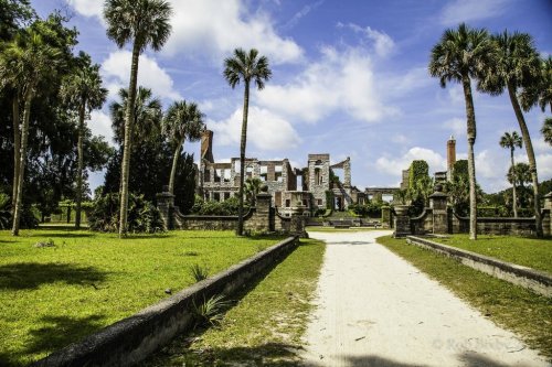 A pathway leads to the ruins of a historic building, surrounded by palm trees and lush greenery under a blue sky.