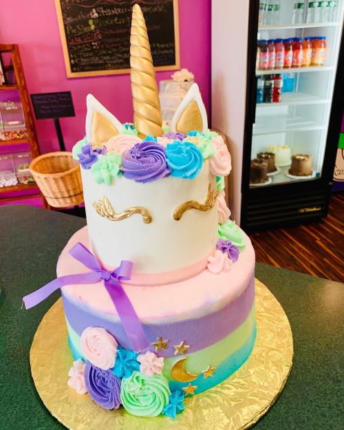 A colorful unicorn-themed cake with a golden horn, pastel frosting, and decorative flowers on a gold cake board.