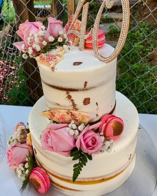 A two-tiered cake decorated with pink roses, macarons, and a "40" topper, set outdoors with a rustic backdrop.
