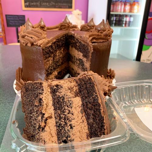A chocolate cake with layers of dark and light chocolate, partially sliced and displayed next to a takeout container.