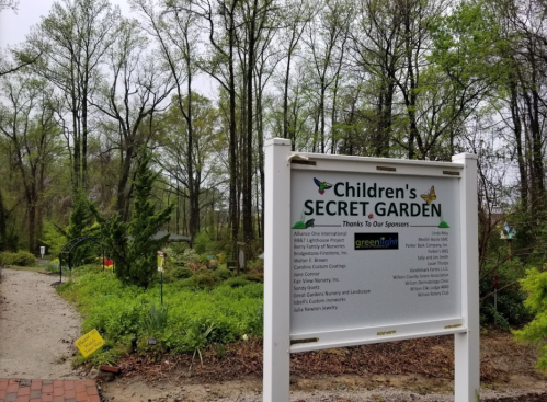 Sign for "Children's Secret Garden" surrounded by greenery, with a list of sponsors and a pathway leading into the garden.