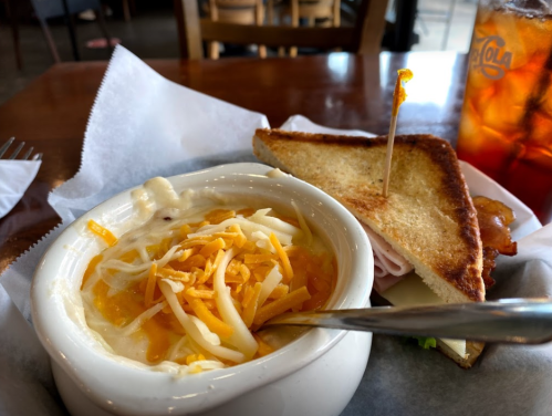 A bowl of creamy soup topped with cheese, alongside a toasted sandwich with ham and lettuce, and a drink in the background.