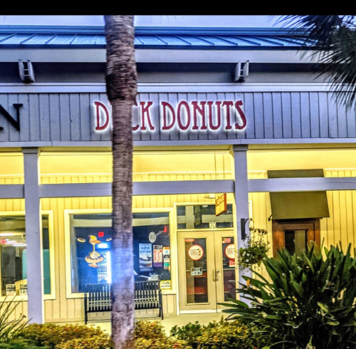 A storefront with the sign "Duck Donuts" illuminated at night, surrounded by palm trees and greenery.