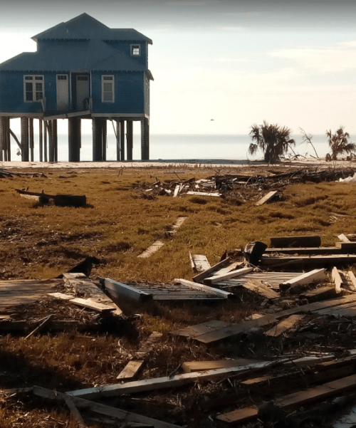 A blue house on stilts near the shore, surrounded by debris and grass, with a calm sea in the background.