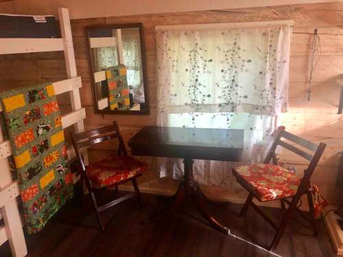 Cozy interior with a small table, two chairs, a mirror, and a colorful quilt on the wall, illuminated by natural light.