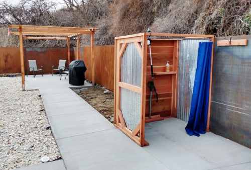 Outdoor shower area with wooden structure, blue curtain, and nearby seating and grill on a paved path.