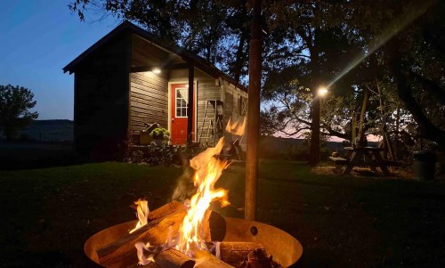 A cozy cabin at dusk, with a warm campfire in the foreground and trees surrounding the peaceful scene.