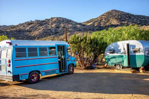 A blue vintage bus parked near a green and silver trailer, surrounded by desert landscape and mountains in the background.