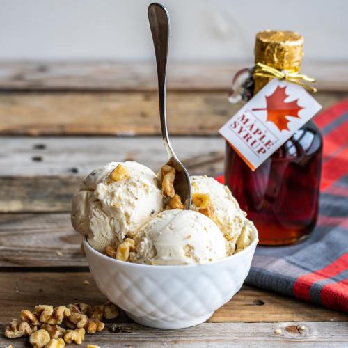 A bowl of vanilla ice cream topped with walnuts, next to a bottle of maple syrup on a wooden table.