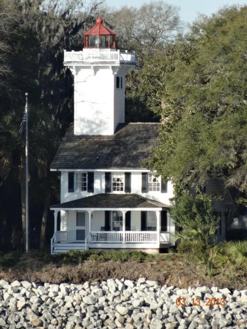 A white lighthouse with a red top stands beside a historic house, surrounded by trees and a rocky shoreline.