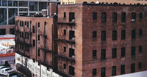 A brick building with fire escapes and air conditioning units, set against a city backdrop.