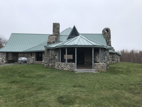 A stone building with a green metal roof, featuring a circular porch and surrounded by grass on a cloudy day.
