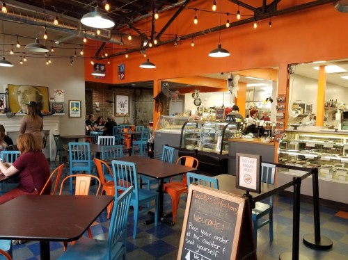 A cozy café interior with colorful chairs, a counter, and a display case, featuring warm lighting and a welcoming atmosphere.