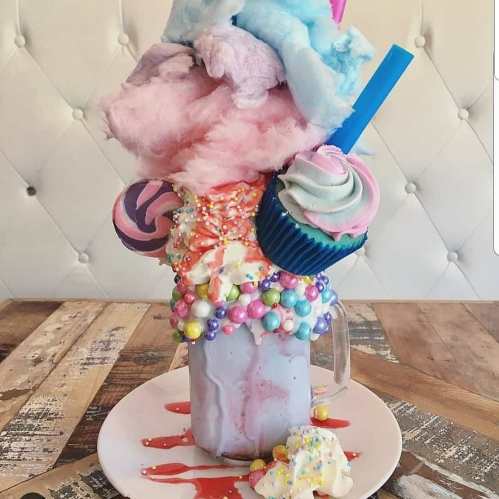 A colorful milkshake topped with cotton candy, cupcakes, and candy, served in a glass on a wooden table.