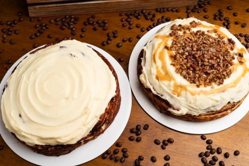 Two round cakes with cream frosting, one topped with pecans and caramel, surrounded by coffee beans on a wooden table.