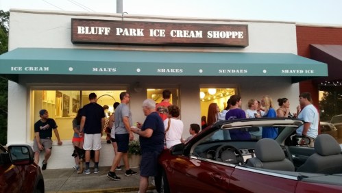 A busy ice cream shop with a crowd outside, enjoying treats on a warm evening. Cars parked nearby.