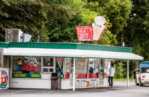 A colorful roadside food stand with a sign reading "Tasty Dip," featuring outdoor seating and a customer ordering.