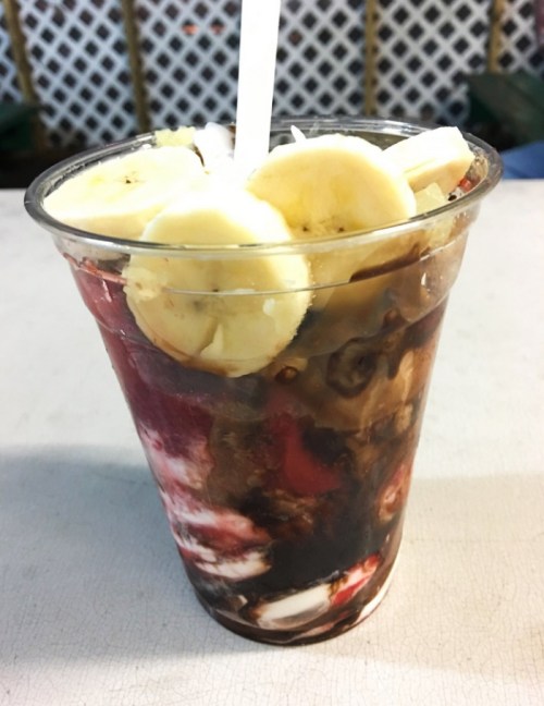 A clear cup filled with layers of chocolate syrup, fruit, and banana slices, topped with a white straw.