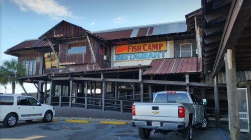 Exterior of Fisht Camp Restaurant with a rustic design, featuring a sign and parked vehicles in front.