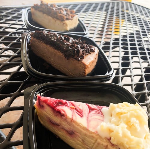 Three slices of cheesecake in black containers: chocolate, strawberry swirl, and white chocolate.