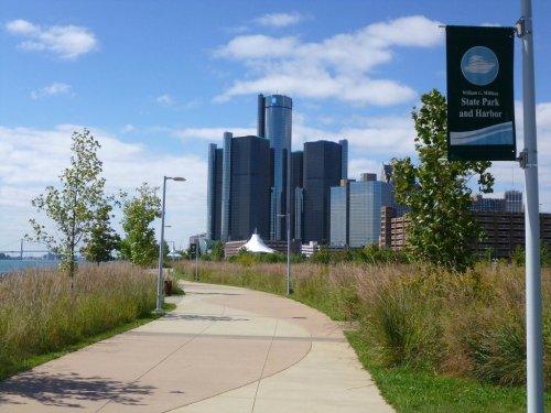 Pathway through greenery leading to a city skyline with modern buildings under a blue sky with clouds.