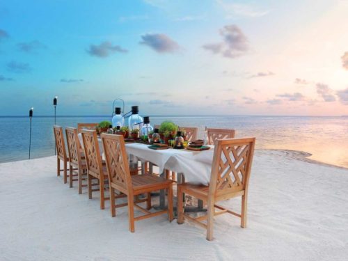 A beautifully set outdoor dining table on a sandy beach at sunset, overlooking the ocean.