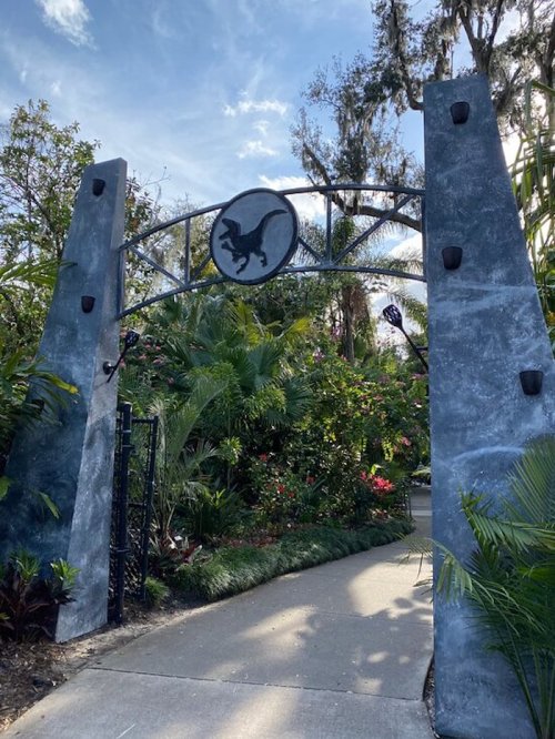 A stone archway with a dinosaur emblem, surrounded by lush greenery and colorful flowers.