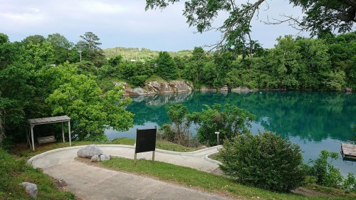A serene lake surrounded by lush greenery, with a path leading down to the water and rocky cliffs in the background.