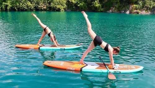 Two women perform yoga poses on paddleboards in a turquoise lake surrounded by greenery.