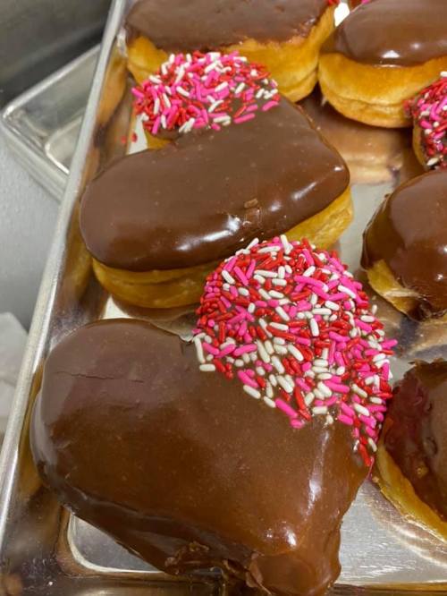 A tray of heart-shaped donuts topped with chocolate icing and colorful sprinkles.
