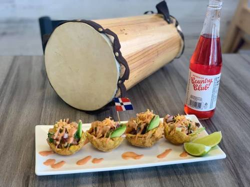 A plate of four tostones topped with seafood, accompanied by a bottle of soda and a wooden drum.