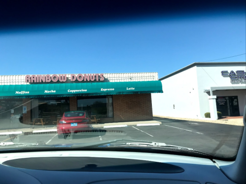 A view of a donut shop named "Rainbow Donuts" with a green awning, parked car in front, and a cash store nearby.