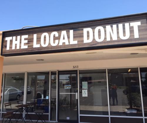 Sign for "The Local Donut" shop with an open door and outdoor seating area. Bright blue sky in the background.