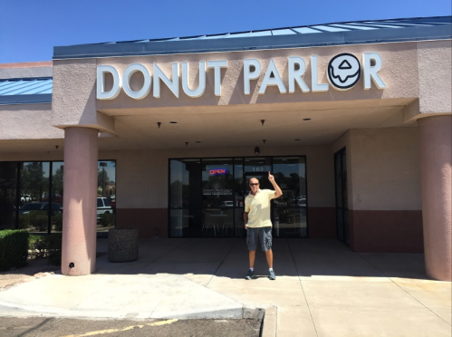 A person stands outside a Donut Parlor, smiling and waving, with a clear blue sky above and an "Open" sign visible.